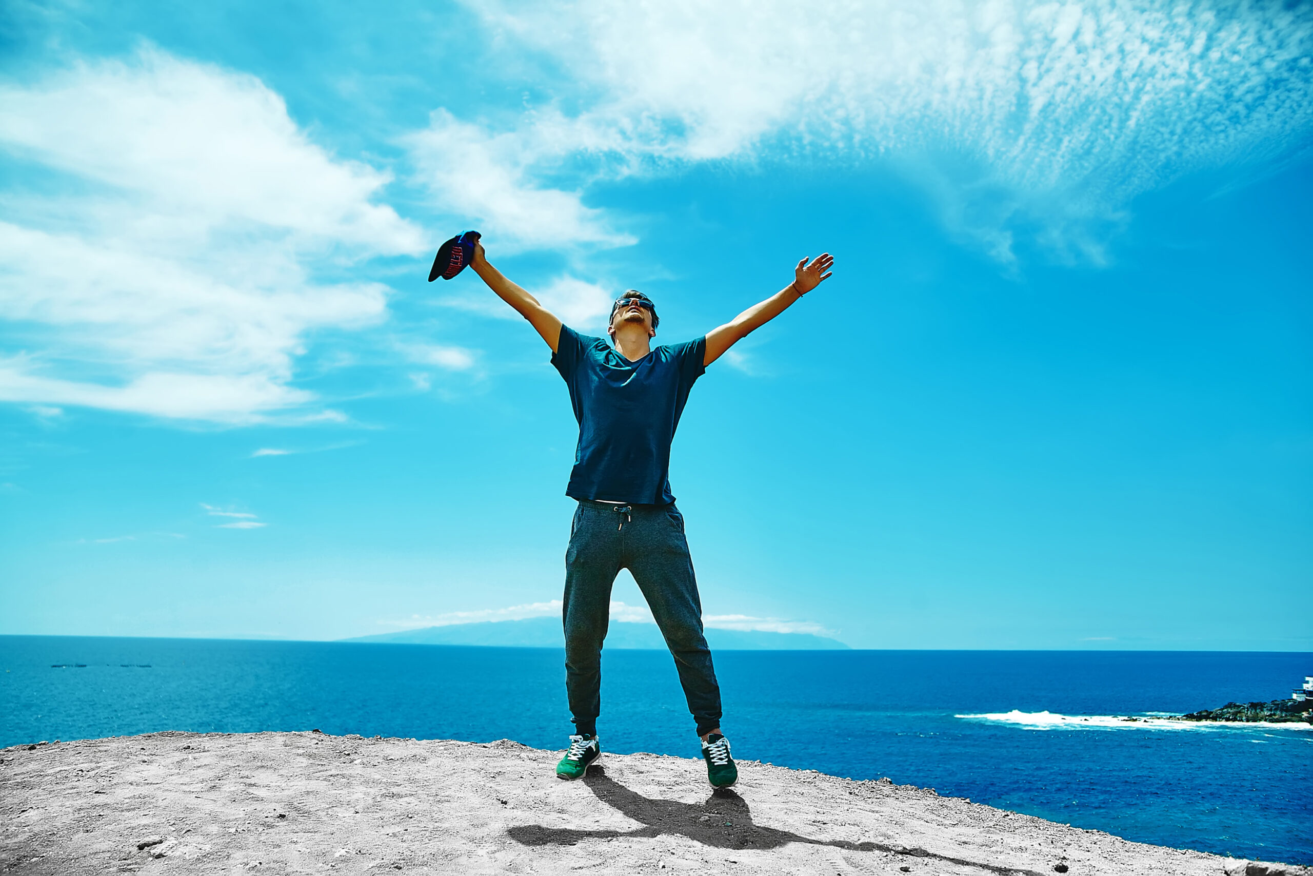 Happy stylish man in casual clothes standing on the cliff of mountain with raised hands to the sun and celebrating success