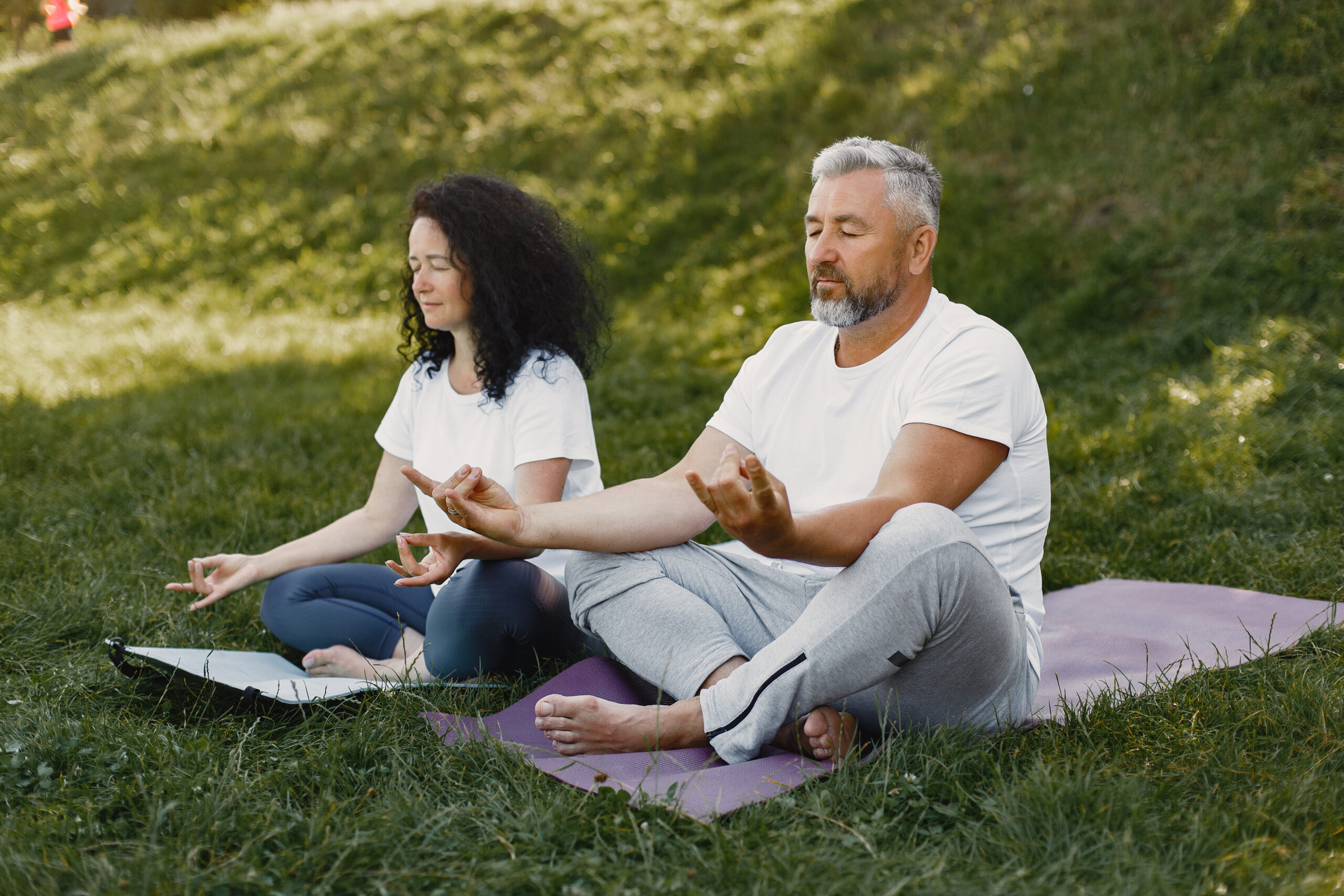 Senior couple is doing yoga outdoors. Stretching in park during sunrise. Brunette in a white t-shirt.